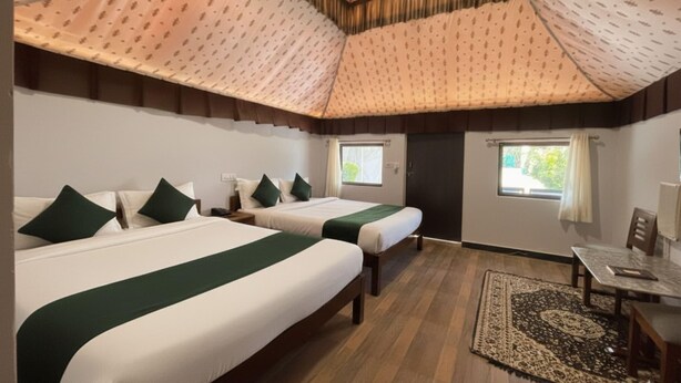 A hotel room with two beds, a small table, and a chair. There are two windows with curtains. - Unity Holiday Resort (Garudeshwar)