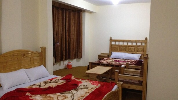 Quarto com duas camas de madeira, uma mesa de cabeceira, uma pequena mesa e uma janela com cortinas.