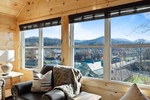 L’intérieur d’un chalet chaleureux, doté d’une grande fenêtre offrant une vue sur une ville et des montagnes.