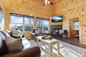 L’intérieur chaleureux d’un chalet avec une cheminée, un canapé, une table et de grandes fenêtres donnant sur le paysage extérieur.