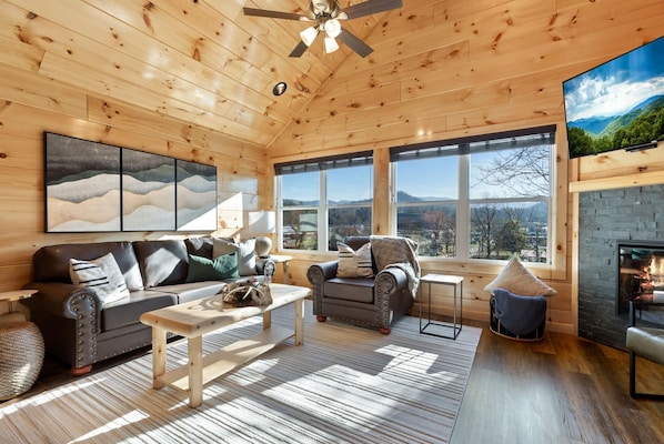 L’intérieur d’un chalet accueillant, doté d’une cheminée, de grandes fenêtres et d’un ventilateur de plafond.