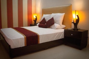 Una cama con ropa blanca y un edredón rojo, flanqueada por dos mesitas de noche con lámparas, en una habitación con papel tapiz a rayas.