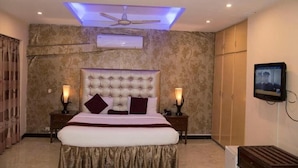 A bedroom with a tufted headboard, a bed with a white and gold bedspread, two bedside lamps, a ceiling fan, an air conditioning unit, a television on the wall, and a wardrobe. - Mall View Hotel Gulberg (Lahore)