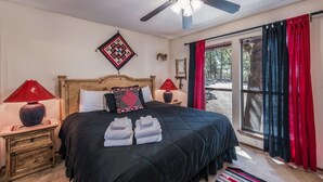 Ein Schlafzimmer mit einem Bett, zwei Nachttischen, einem Deckenventilator, einem Fenster mit Vorhängen und Blick ins Freie.