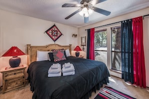 A bedroom with a bed, two nightstands, a ceiling fan, a window with curtains, and a view of the outdoors. - Cowboys Indians And Outlaws By Vtrips (Ruidoso)