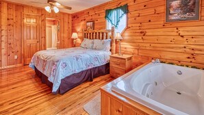 Ein Schlafzimmer einer Holzhütte mit einem Bett, einem Nachttisch, einer Lampe, einem Fenster mit Vorhängen, einem Deckenventilator und einer Badewanne.