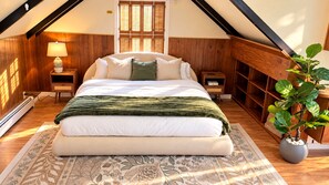 Un dormitorio con una cama grande, paredes de madera, una ventana con contraventanas, una mesita de noche y una lámpara, además de una planta en maceta.
