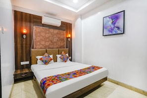 A hotel room with a bed, a headboard with a patterned design, an air conditioning unit, a wall-mounted artwork, and a bedside table with a lamp. - Fabhotel Sai Plaza Executive (Chhatrapati Sambhajinagar)