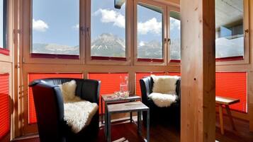 Ein Zimmer mit Holzvertäfelung, zwei schwarze Ledersessel mit weißen Felldecken, ein kleiner Tisch mit einem Glas und Blick auf Berge durch große Fenster.