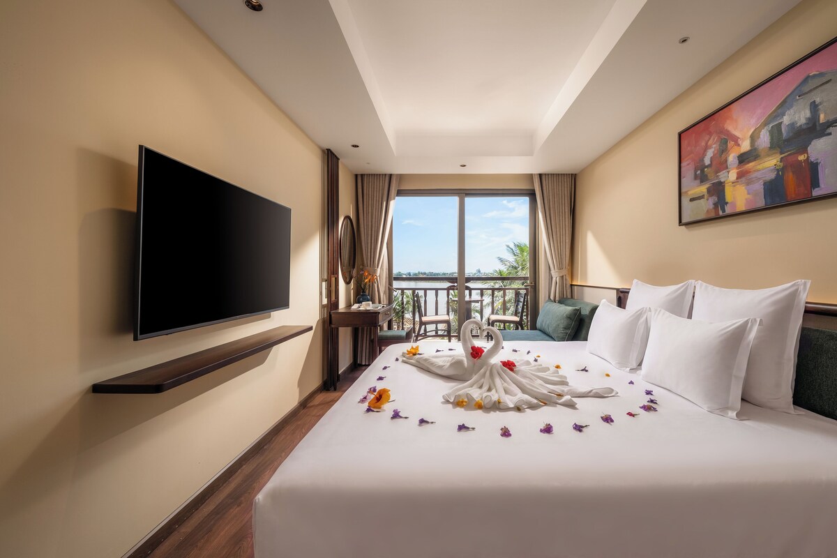 A hotel room with a large bed, a flat-screen TV mounted on the wall, a balcony with a view, and a painting on the wall.