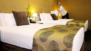 Twee bedden met witte en bruine beddengoed, een nachtkastje met een lamp en een plant in een hotelkamer.