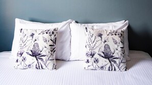 Een slaapkamer met een bed, twee kussens met bloemenmotieven en twee ingelijste botanische prints aan de muur.