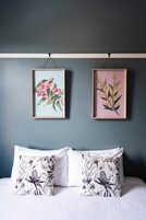 Un dormitorio con cama, dos almohadas estampadas con motivos florales y dos láminas botánicas enmarcadas en la pared.