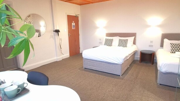 A hotel room with a wooden ceiling, a bed with pillows, a bedside table, a chair, and a round table with cups and a tray. - The Elmsleigh Suite At Claremont Hotel (Nottingham)