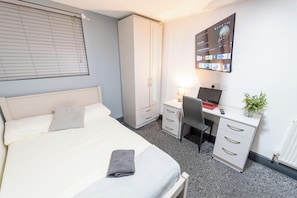 Quarto com cama, escrivaninha, cadeira, guarda-roupa e televisão presa à parede.