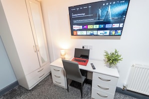 Quarto de hotel com escrivaninha, laptop, controle remoto, planta em vaso e televisão na parede.