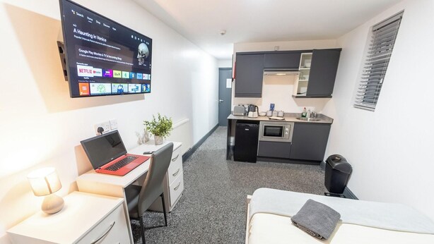 A compact hotel room with a bed, a desk with a laptop, a small kitchenette, and a TV mounted on the wall displaying a movie. - Charming Studio In Birmingham (Birmingham)