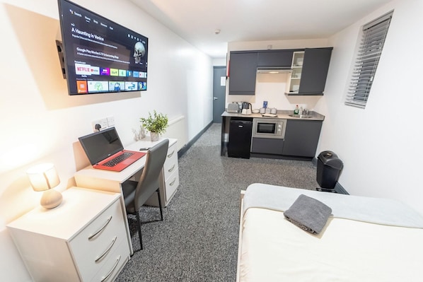Quarto de hotel compacto com cama, escrivaninha com laptop, pequeno espaço de cozinha e televisão na parede exibindo um filme.
