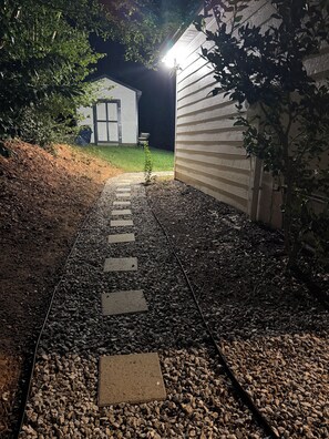 Ein Weg führt zu einem Haus, über dem eine Lichtquelle hängt, umgeben von Kies und Vegetation.
