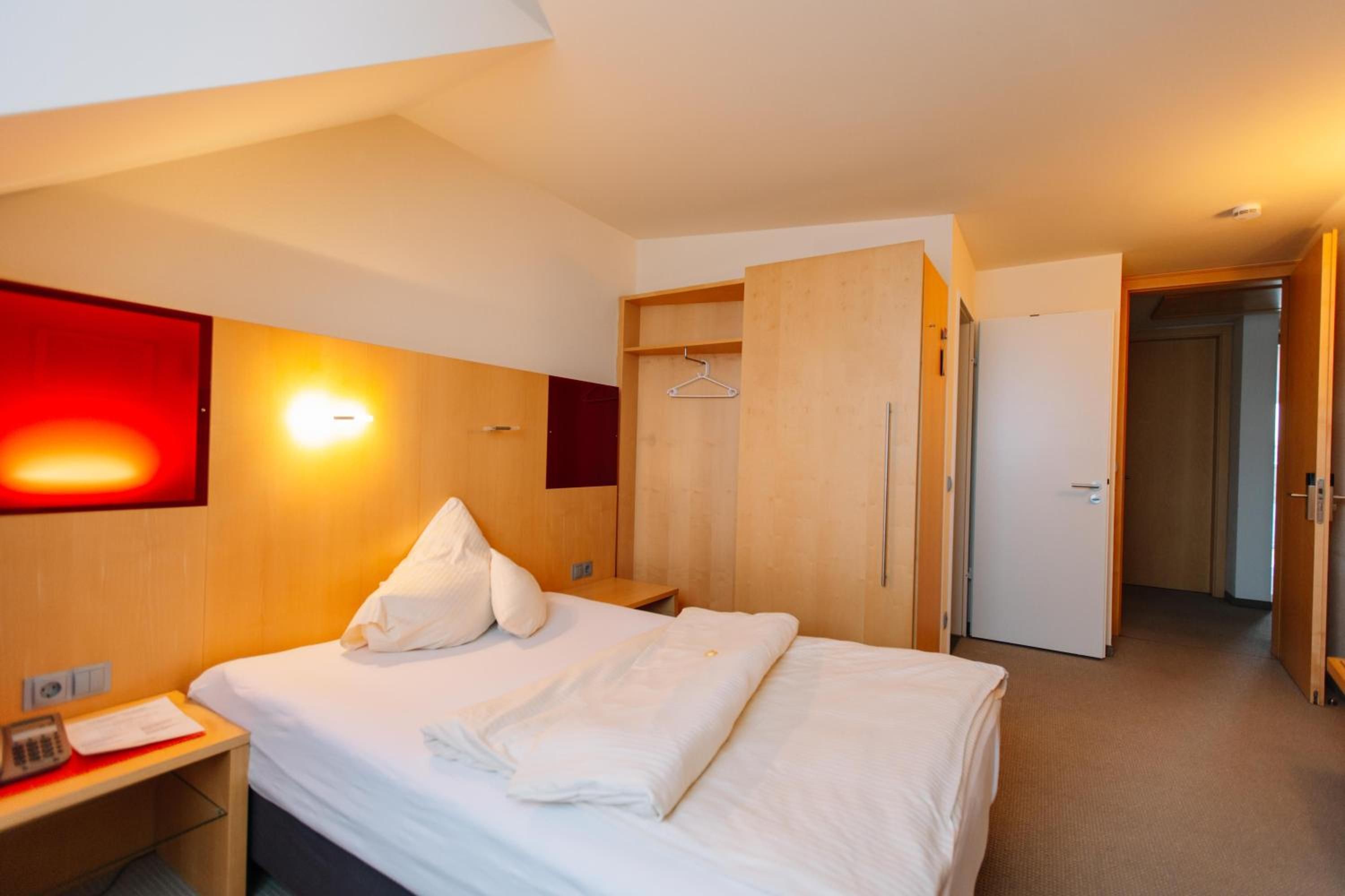 A hotel room with a bed, a desk with a telephone, a wardrobe, and a red wall with a light fixture.