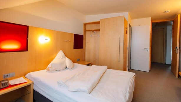 A hotel room with a bed, a desk with a telephone, a wardrobe, and a red wall with a light fixture. - Landhotel Gasthof Drexler Gbr (Fuerstenfeldbruck)