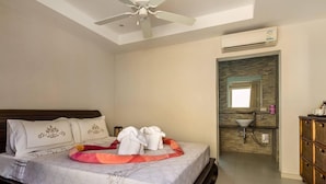 Een slaapkamer met een bed, een plafondventilator, een airconditioner, een badkamer met een wastafel en spiegel, en een houten dressoir.