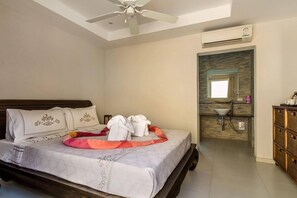 Un dormitorio con cama, ventilador de techo, unidad de aire acondicionado, baño con lavamanos y espejo, y cómoda de madera.
