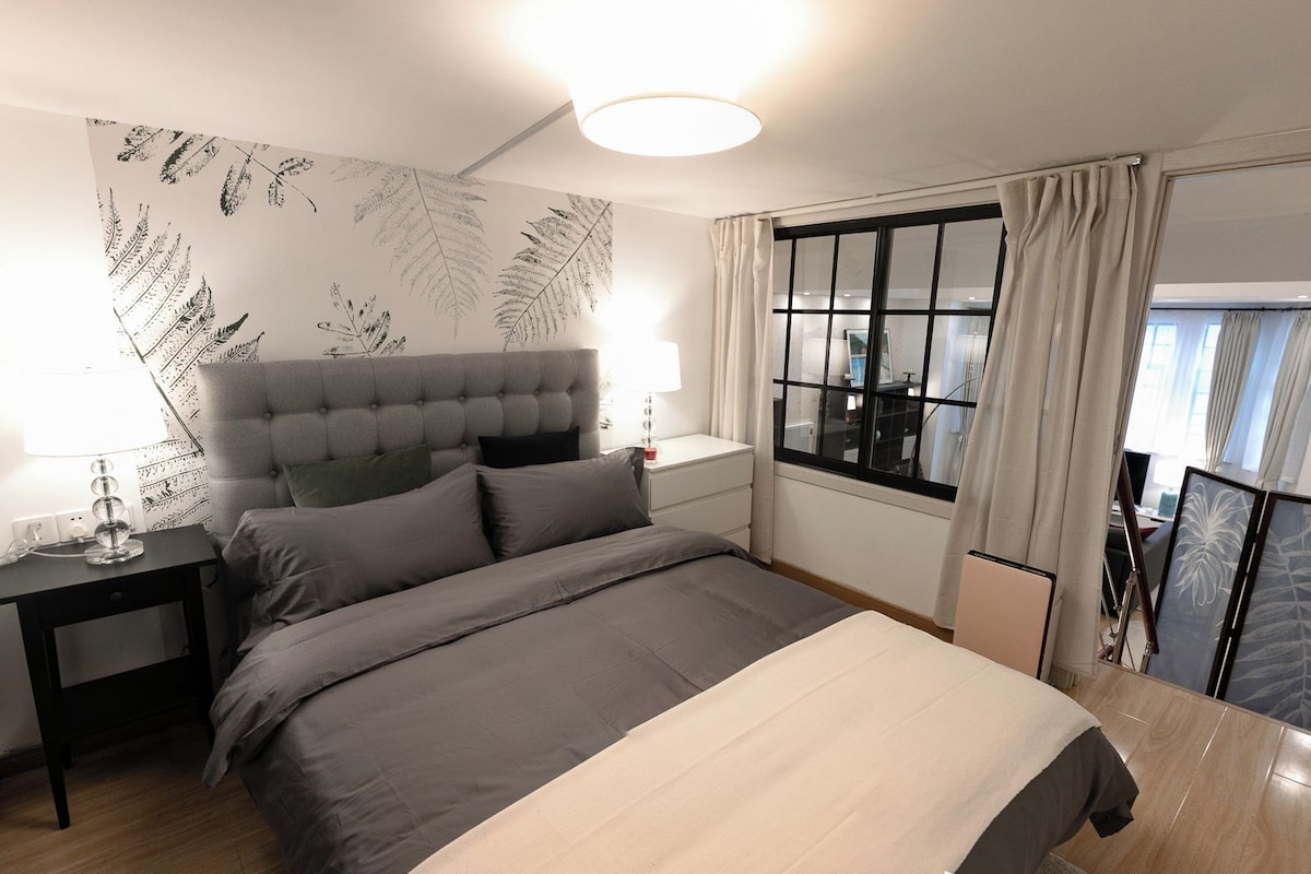 A modern bedroom with a large bed, bedside tables, a nightstand, a window with curtains, and a view of the outside.