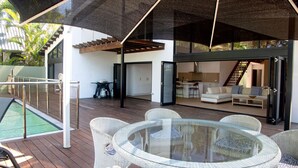 An outdoor patio with a glass table and wicker chairs, a pool, and a covered area with a staircase leading to an upper level. - The Barrington- Ocean Front Views (Wongaling Beach)