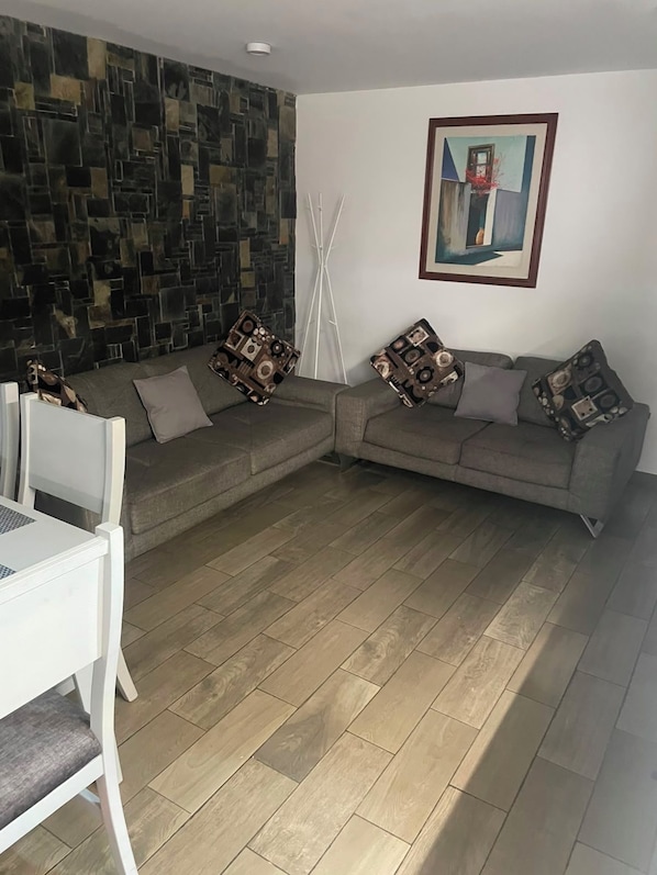 Uma sala de estar com sofá de canto, uma mesa branca e parede estampada.