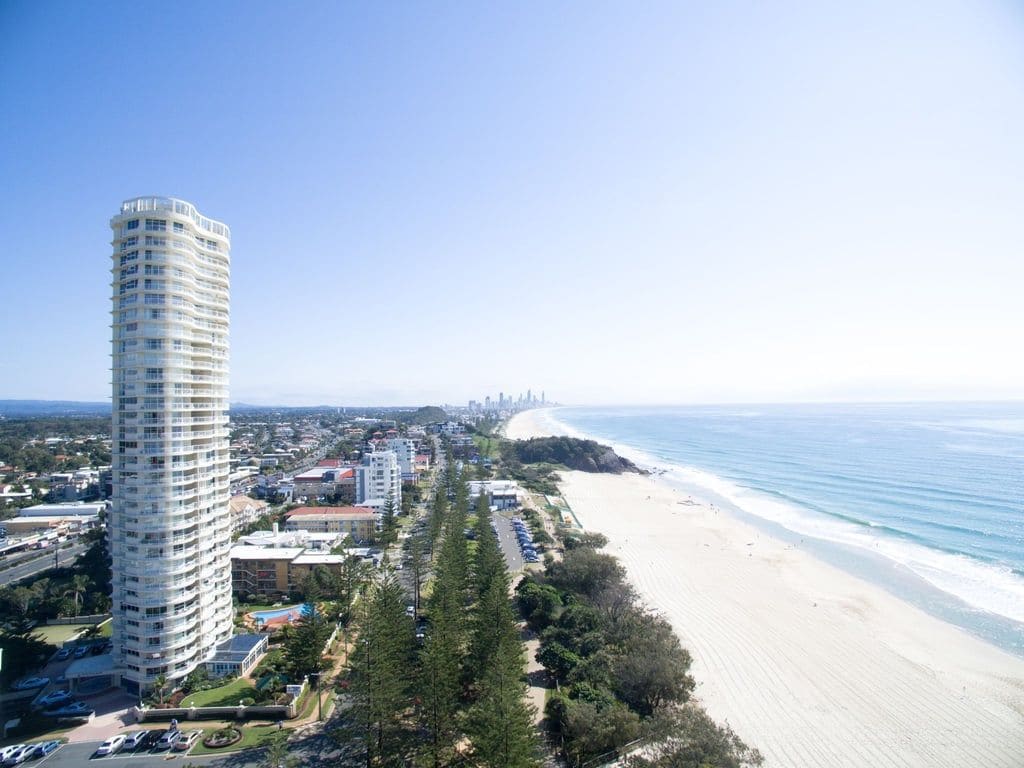 Un paisaje urbano costero con un hotel moderno imponente, playa y una silueta de la ciudad en la distancia.
