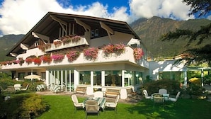 A chalet with a balcony adorned with flowers, a covered outdoor seating area, and a mountain backdrop. - Palmengarten Hotel Gstv R (Lagundo)