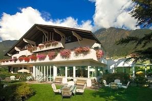 A chalet with a balcony adorned with flowers, a covered outdoor seating area, and a mountain backdrop. - Palmengarten Hotel Gstv R (Lagundo)