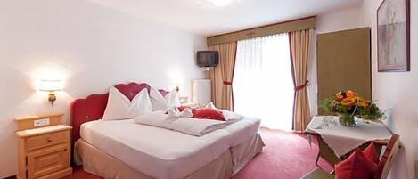 A hotel room with a bed, bedside table, lamp, TV, and a small table with a vase of flowers.