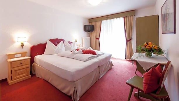 A hotel room with a bed, bedside table, lamp, TV, and a small table with a vase of flowers. - Palmengarten Hotel Gstv R (Lagundo)