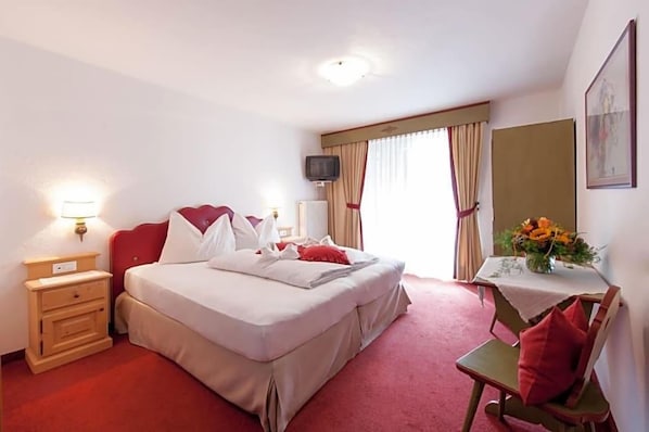 A hotel room with a bed, bedside table, lamp, TV, and a small table with a vase of flowers. - Palmengarten Hotel Gstv R (Lagundo)