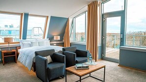 Habitación de hotel con una cama, dos sillones, una mesita de centro y grandes ventanales con vistas a la ciudad.