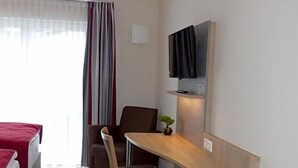 A hotel room with a bed, a desk with a chair, a television mounted on the wall, and a window with curtains. - Ladiges Gasthof (Holm)