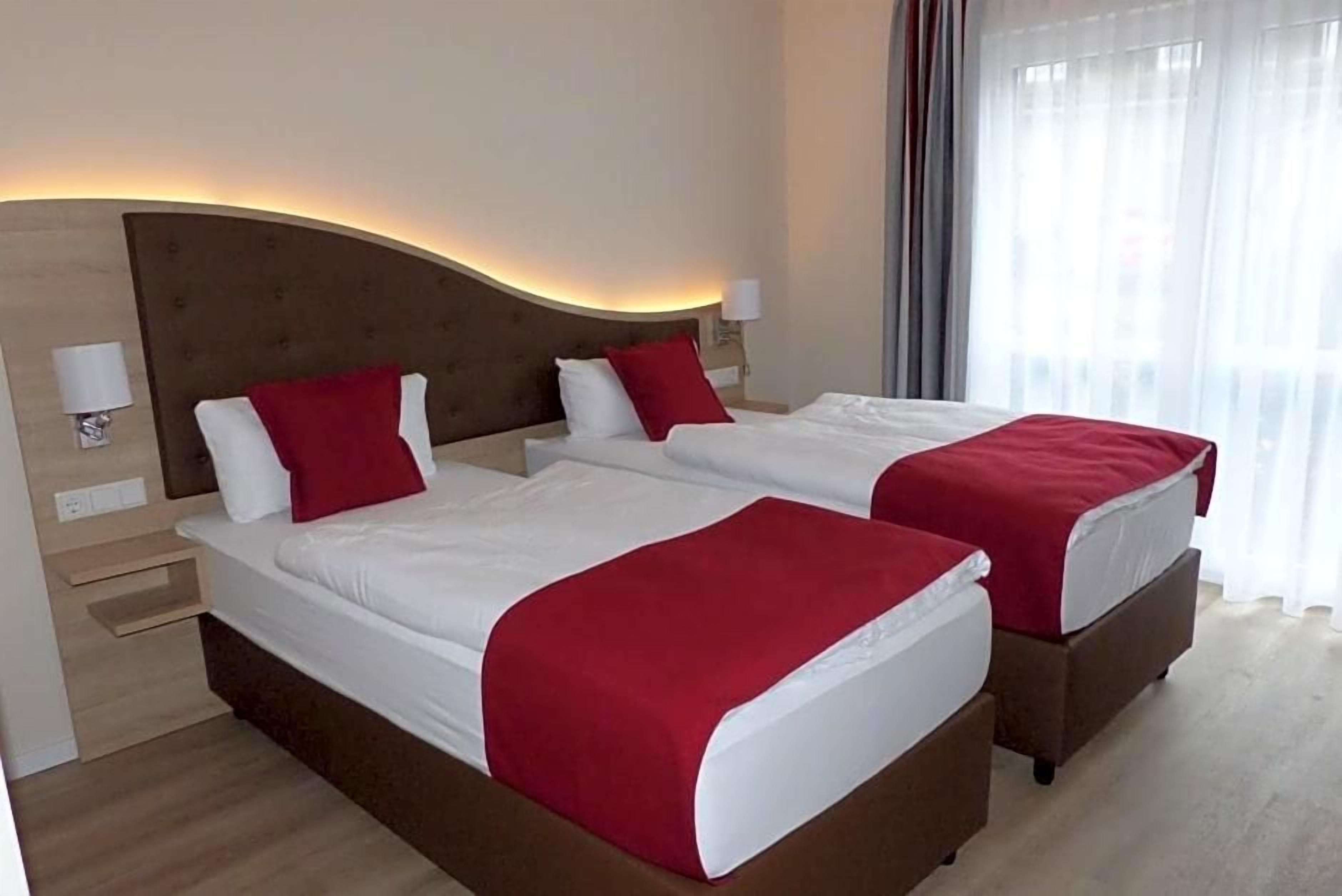 A hotel room with two beds, each with a red bedspread, a wooden headboard, and a bedside table with a lamp.