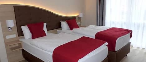 Habitación de hotel con dos camas, cada una con colcha roja, cabecero de madera y mesita de noche con lámpara.