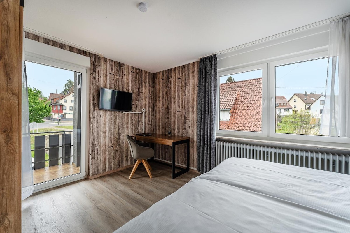 A bedroom with a bed, a desk, a chair, a television, and a window with a view of houses.