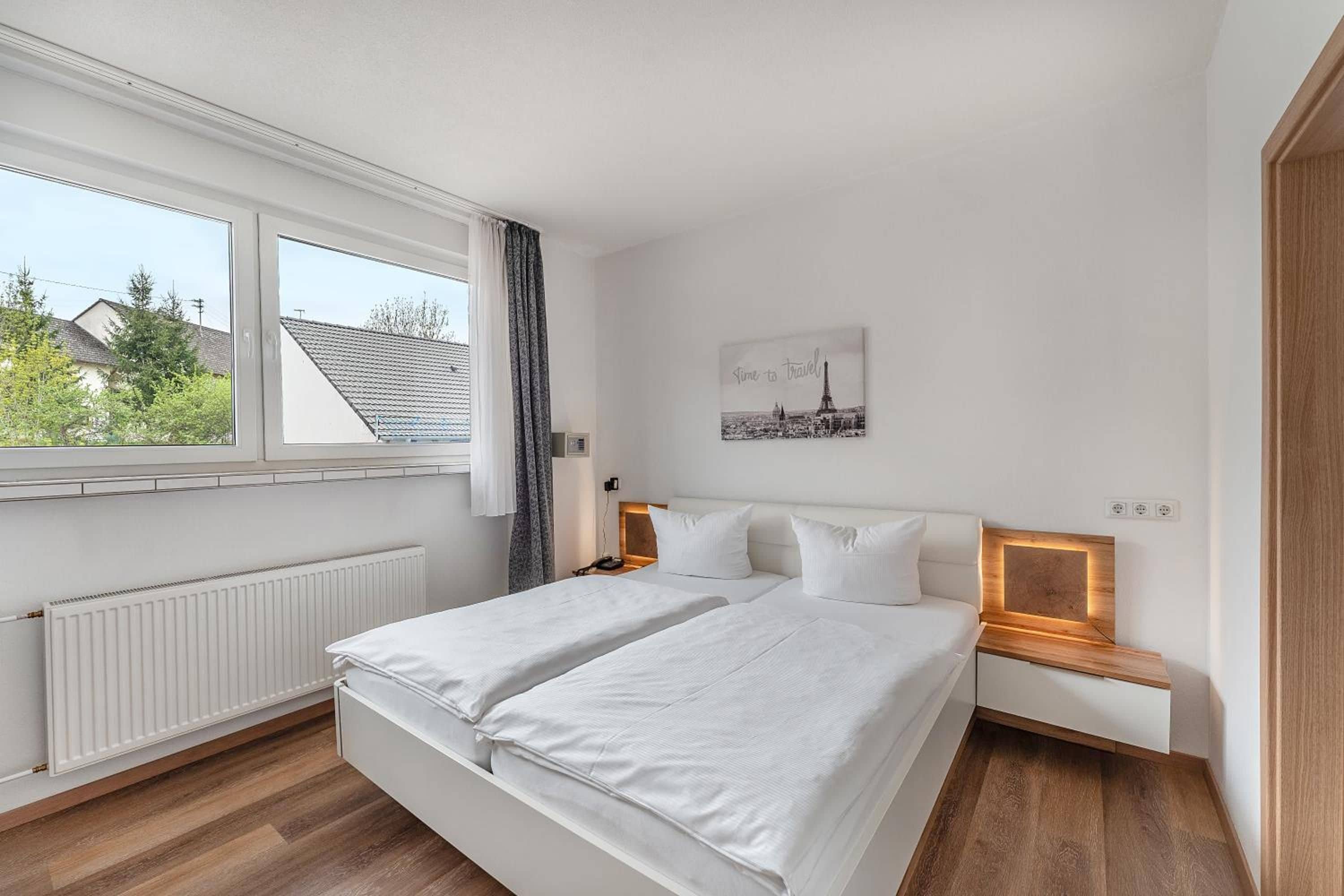 A modern bedroom with a large bed, a bedside table, a nightstand, a window with a view of houses, and a wall-mounted picture.