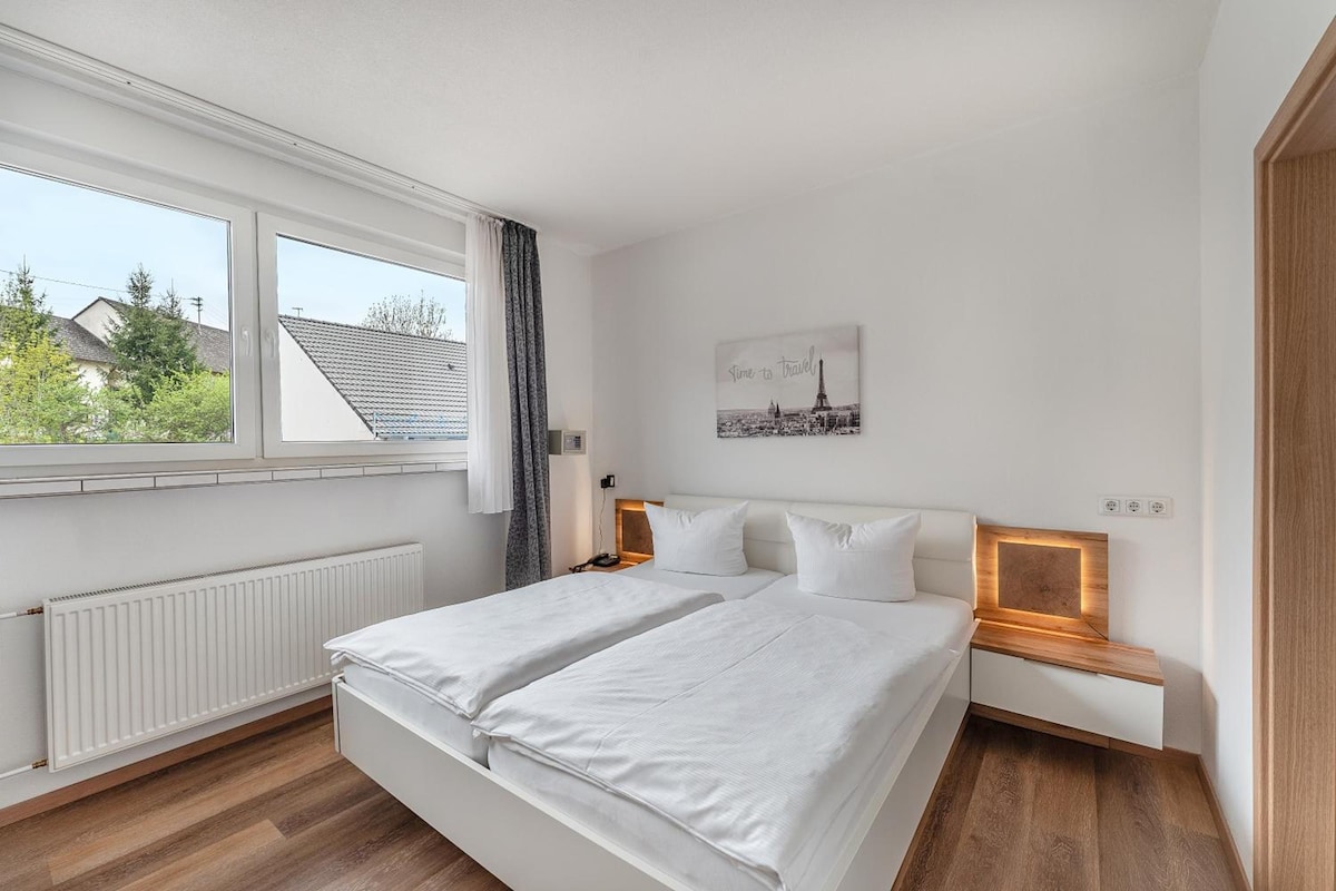 A modern bedroom with a large bed, a bedside table, a nightstand, a window with a view of houses, and a wall-mounted picture.