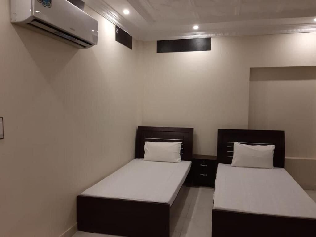 A hotel room with two single beds, a small bedside table, a wall-mounted air conditioner, and a plain wall.
