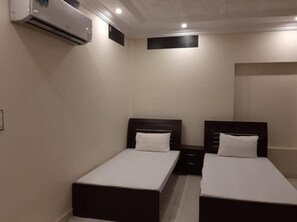 Une chambre d’hôtel avec deux lits simples, une petite table de chevet, un climatiseur fixé au mur et un mur sobre.