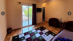 Quarto de hotel com piso de madeira, tapete acolchoado, cama, cadeira, TV fixada na parede e varanda com vista para edifícios.