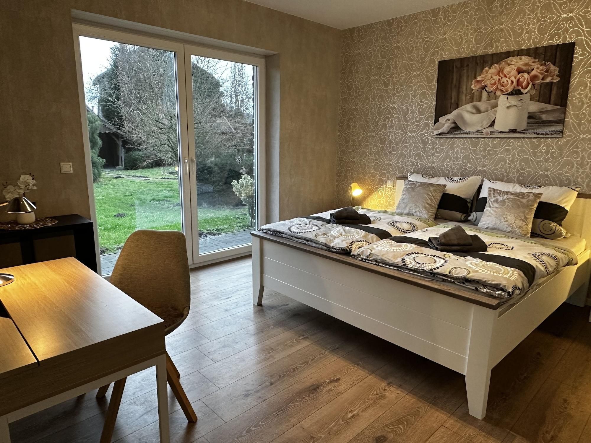 Un dormitorio con cama, escritorio y silla. Se ve un ventanal con vistas a árboles y césped.