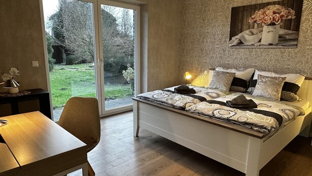 A bedroom with a bed, a desk, and a chair. There is a window with a view of trees and a lawn. - Ferienhaus Am Gla Ck (Bad Zwischenahn)