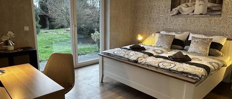 Un dormitorio con cama, escritorio y silla. Se ve un ventanal con vistas a árboles y césped.