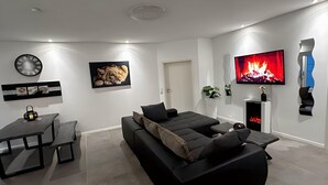 A modern living room with a black sofa, a flat-screen TV, a fireplace, and a dining area with a table and chairs. - Ferienhaus Am Gla Ck (Bad Zwischenahn)